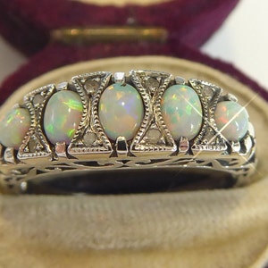 Könnte beinhalten: Ein silberner Ring mit fünf ovalen Opalen mit schillernden Farben. Die Opale sind in einem filigranen Design mit kleinen Akzentsteinen gefasst. Der Ring wird in einer Samt-Schmuckschatulle präsentiert.