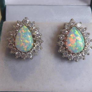 May include: A pair of silver earrings featuring pear-shaped opals set in a cluster of small clear stones.