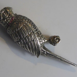 May include: A silver parrot brooch with red gemstone accents. The parrot is detailed with feathers and has a long tail.