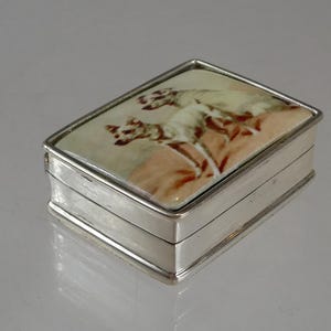 May include: A rectangular silver box with a hinged lid featuring an inset ceramic panel. The panel displays a painted illustration of two white dogs standing on a tan surface. The metal box has a polished finish and a simple, clean-lined design.