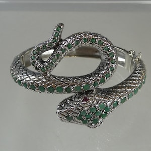 May include: A silver-toned snake bracelet with green and red gemstone accents. The bracelet is designed to wrap around the wrist, with the snake's head and tail meeting. The snake's body is textured, and the gemstones are set in the design.