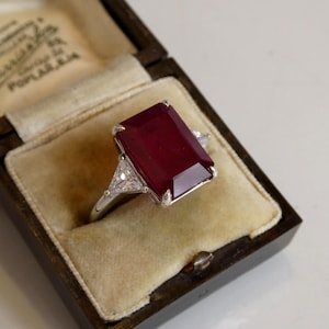 Könnte beinhalten: Ein rechteckiger Ring mit tiefrotem Edelstein und zwei dreieckigen Akzentsteinen. Der Ring ist in einem silbernen Band gefasst und wird in einer Vintage-Schmuckschatulle aus Holz mit cremefarbenem Samtinterieur präsentiert.