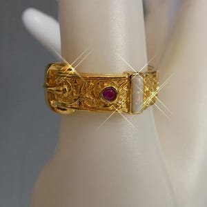 May include: A gold-colored ring with intricate floral patterns and two inset gemstones. One gemstone is a small, round, red ruby, and the other is a rectangular opal. The ring has a buckle design.