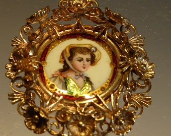 May include: A gold-toned pendant with a delicate floral design. The pendant features a round portrait of a young woman with a white bonnet and a green and gold dress. The portrait is set in a red and gold frame.