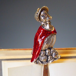 Puede incluir: Marcapáginas de plata con un pato que lleva una capa roja y un gorro. El pato sostiene un grupo de patitos.