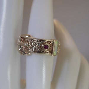 Könnte beinhalten: Ein goldener Ring mit einem detaillierten Schnallenmuster, verziert mit einem kleinen roten Edelstein und einem größeren weißen Opal.