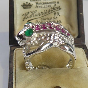 Puede incluir: Un anillo de plata en forma de rana con ojos verde esmeralda y piedras de rubí rojo engastadas en el cuerpo. El anillo está en una caja de terciopelo marrón con una etiqueta que dice "H. Harris & Son, Relojeros y Joyeros, 89, Chrisp St., Pop 14."