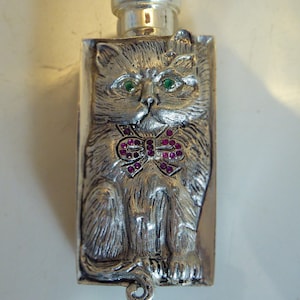 Op de afbeelding: Zilveren parfumfles met een kattenontwerp. De kat heeft groene ogen en een roze strik met strass.