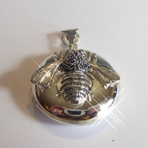 May include: A silver pendant shaped like a bee, with detailed wings and body. The bee is mounted on a round, polished base. The pendant has a loop at the top for a chain, and is highly reflective.