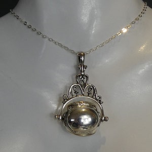 May include: A silver pendant necklace with an elaborate design. The pendant has a rotating locket-style compartment and hangs from a fine chain. The design incorporates decorative elements and a detailed pattern. The necklace is a classic piece.