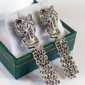 May include: A pair of silver dangle earrings featuring a wolf head design. The wolf heads are covered in black marcasite stones and have red ruby eyes. The dangle portion of the earrings is made of silver and is adorned with black marcasite stones, green emerald stones, and red ruby stones.
