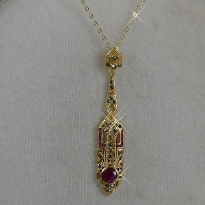 May include: A gold pendant necklace featuring an elongated, geometric design with intricate metalwork, small dark stones, rectangular pink accents, and a single round, deep pink gemstone at the base, suspended from a delicate gold chain.