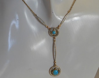 Op de afbeelding: Een gouden ketting met twee turquoise stenen gezet in witte emaille cirkels. De ketting heeft een delicate ketting en een Y-vormig ontwerp.