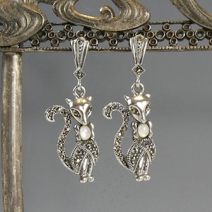 Puede incluir: Un par de pendientes colgantes de plata con un diseño de gato con detalles de marcasita y una perla blanca en el centro del pecho de cada gato.