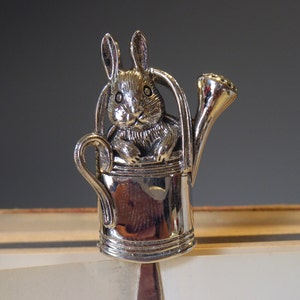 Puede incluir: Figura de conejo de plata sentada dentro de una regadera de plata con un pico. El conejo sostiene una pequeña flor en su pata.