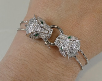May include: A silver bracelet with two panther heads, each set with diamonds and green emerald eyes. The panther heads are connected by a silver chain.
