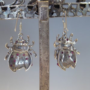 May include: A pair of silver ladybug earrings with red gemstone accents. The earrings are hanging from a silver stand with the text "Catherine Popesco France" on it.