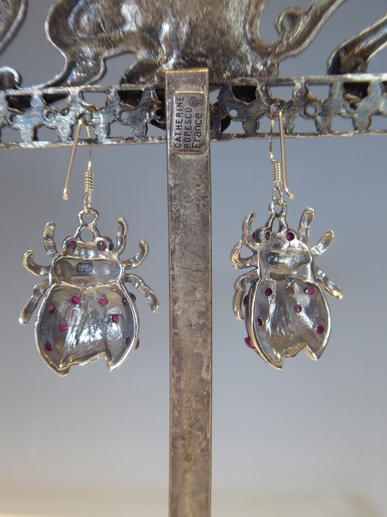 May include: A pair of silver ladybug earrings with red gemstone accents. The earrings are hanging from a silver stand with the text "Catherine Popesco France" on it.