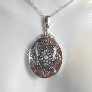 Puede incluir: Un collar con un dije de locket ovalado de plata, con una cadena delicada. El locket tiene un diseño ornamentado con una piedra de ópalo blanco en el centro.