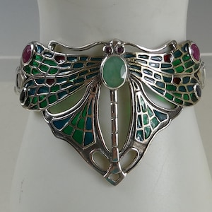 May include: A silver cuff bracelet shaped like a dragonfly, featuring green and blue enamel wing patterns, a central oval light green gemstone, and small round red gemstones set at the wing tips and near the head of the dragonfly design.