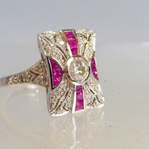 Può includere: Un anello vintage con un grande diamante rotondo e chiaro circondato da diamanti più piccoli e rubini rossi. L'anello è incastonato in un design rettangolare e filigranato.