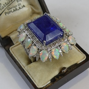 May include: A large, blue gemstone ring set in silver with a halo of white opals and diamonds. The ring is in a small, wooden box with a label that reads "Harris & Son, Watchmakers & Jewellers, 89, Chrisp St, Poplar, E.14."