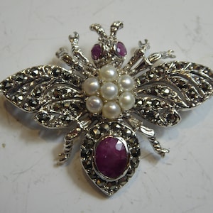 Puede incluir: Un broche de abeja de plata con un grupo de perlas blancas en el centro, dos piedras de rubí rojo en la cabeza y una piedra de rubí rojo más grande en el abdomen. La abeja está rodeada por un diseño detallado de piedras de pirita.