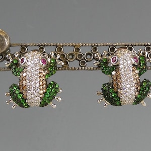 May include: A pair of frog-shaped earrings. Each frog is adorned with white, brown, and green gemstones, with ruby eyes. The earrings are attached to a silver bar with decorative elements.