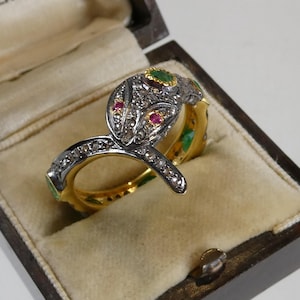 Op de afbeelding: Een gedetailleerde slangvormige ring met een zilveren kop en gouden band, bezet met kleine diamanten, robijnen en smaragden. De ring wordt gepresenteerd in een houten sieradendoos met een beige interieur.