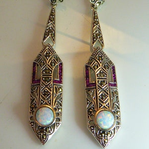 May include: A pair of silver dangle earrings featuring a white opal gemstone and pink accents. The earrings have a filigree design with a geometric pattern.