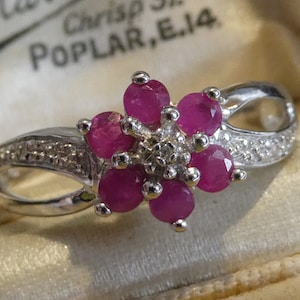 May include: A silver ring with a flower design featuring pink gemstones and smaller white gemstones. The ring is in a white box with the text "Crisp St. POPLAR, E.14".