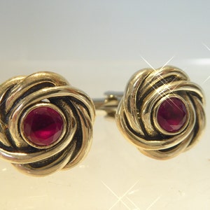 May include: A pair of gold cufflinks with a red gemstone in the centre of each. The cufflinks are in the shape of a knot.