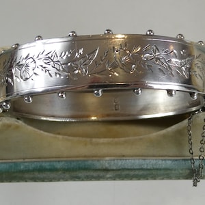 Puede incluir: Un brazalete rígido articulado de tono plateado con un patrón floral grabado en el centro. Los bordes del brazalete están decorados con pequeñas esferas metálicas espaciadas uniformemente. Una fina cadena de seguridad está unida al cierre lateral.