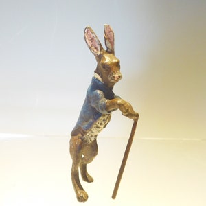 May include: A small bronze figurine of a rabbit wearing a blue jacket and holding a walking stick. The rabbit has pink ears and brown fur.
