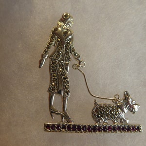 Op de afbeelding: Zilverkleurige broche met een vrouw die een hond aan de lijn uitlaat. De vrouw draagt een jas en schoenen. De hond is een Scottish Terrier. De basis van de broche is bezet met kleine, rode edelstenen.