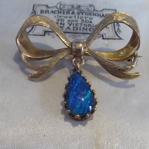 May include: A gold bow brooch with a teardrop-shaped blue opal pendant. The bow is textured and has a hammered finish.