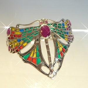 Pode incluir: Um bracelete de prata em forma de libélula com detalhes em esmalte colorido e uma grande pedra de rubi no centro.