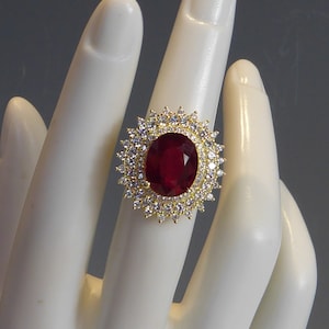 以下が含まれることがあります： 大きな楕円形の赤い宝石が、小さな透明な宝石で囲まれた金の指輪