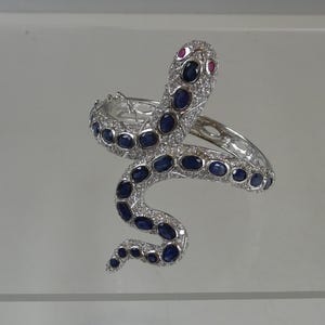 May include: A silver snake bracelet with blue and clear gemstones. The snake's head features two red gemstones for eyes. The body of the snake is adorned with oval-shaped blue gemstones and clear gemstones.