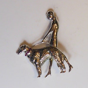 Op de afbeelding: Een zilveren broche met daarop een vrouw in rijkkleding die een windhond uitlaat. De vrouw draagt een hoed en de hond heeft een rode robijn-halsband.