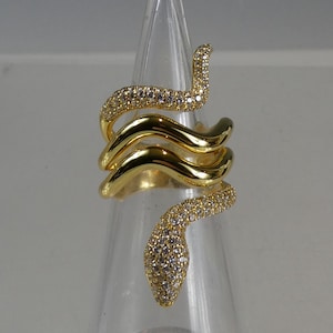 Op de afbeelding: Een goudkleurige ring in de vorm van een slang, waarbij het lichaam van de slang om de vinger is gewikkeld. De kop van de slang en een deel van zijn lichaam zijn bezet met kleine, heldere edelstenen. De ring wordt getoond op een transparante, kegelvormige standaard.