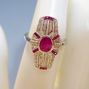 May include: A silver ring with a large oval ruby surrounded by smaller rubies and diamonds. The ring has a geometric design.
