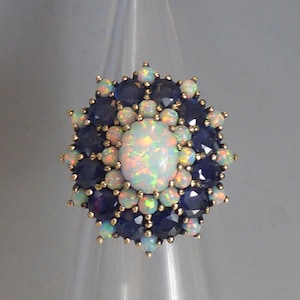 Pode incluir: Um anel de ouro com uma grande opala branca cercada por pequenos safiras azuis e opalas brancas.