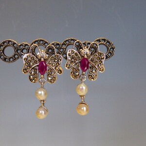 May include: A pair of butterfly-shaped earrings. Each earring features a central ruby gemstone, surrounded by small, sparkling stones. Below the butterfly design, two dangling pearls add a touch of elegance. The earrings are set against a neutral background.