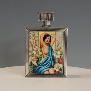 Può includere: Un flacone di profumo in argento con un design rettangolare. Il flacone presenta un'immagine colorata di una donna con un vestito blu in piedi in un giardino.