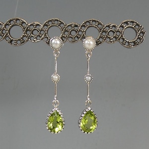 May include: Silver drop earrings with pear-shaped, light green peridot gemstones. Each earring has a small pearl at the top and a smaller pearl down the silver chain. The earrings are displayed on a decorative silver bar with a repeating pattern.