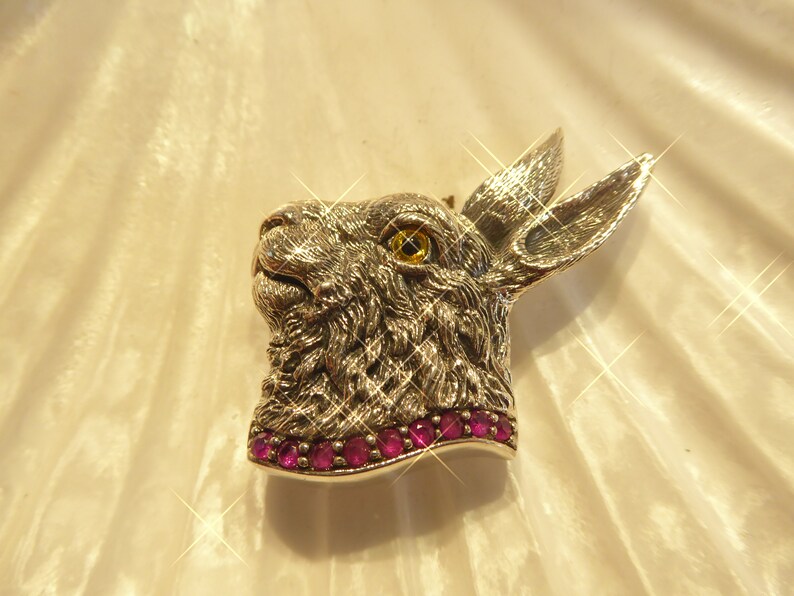May include: A silver rabbit head brooch with a yellow eye and a row of pink gemstones around the neck.