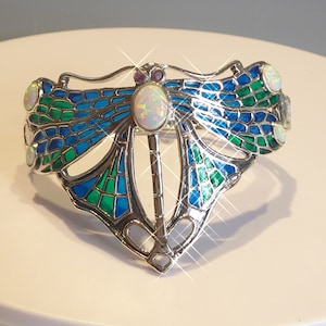 May include: A silver dragonfly bracelet with blue and green enamel, featuring an opal gemstone in the center and smaller opals on the wings.