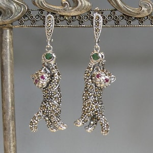 Puede incluir: Un par de pendientes de plata con diseño de pantera. Los pendientes están adornados con piedras de marcasita y tienen piedras preciosas rojas y verdes.