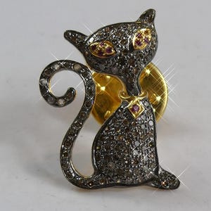 Può includere: Spilla a forma di gatto nero con occhi di rubino e corpo tempestato di diamanti. Il gatto è seduto con la coda arricciata. La spilla ha un retro dorato e un piccolo rubino sul colletto.